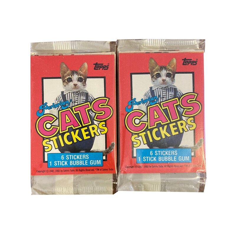 May include: Two packs of Topps Perlerian Cats Stickers, each featuring a cartoon cat wearing a blue and white striped shirt and a red bow tie. Each pack contains 6 stickers and 1 stick of bubble gum. The text on the packs reads "Topps Perlerian Cats Stickers" and "6 Stickers 1 Stick Bubble Gum".