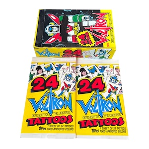 May include: A yellow box and two packs of Voltron Defender of the Universe tattoos. The box features a Voltron robot design. The packs have the number 24 in large red font, with the Voltron logo and the text "Tattoos".