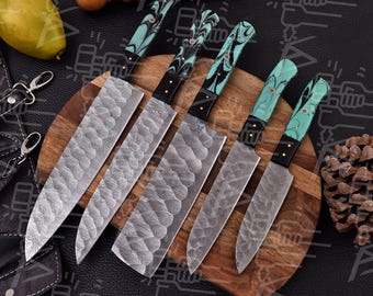 May include: A collection of six kitchen knives with textured silver blades and turquoise, black, and white patterned handles. The knives are displayed on a wooden chopping board, with a wooden mallet and a pear in the background. The knives are for food preparation.