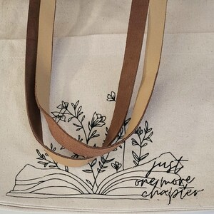 May include: Cream tote bag with brown and tan leather straps. The bag has a black line drawing of an open book with floral accents and the text "just one more chapter."