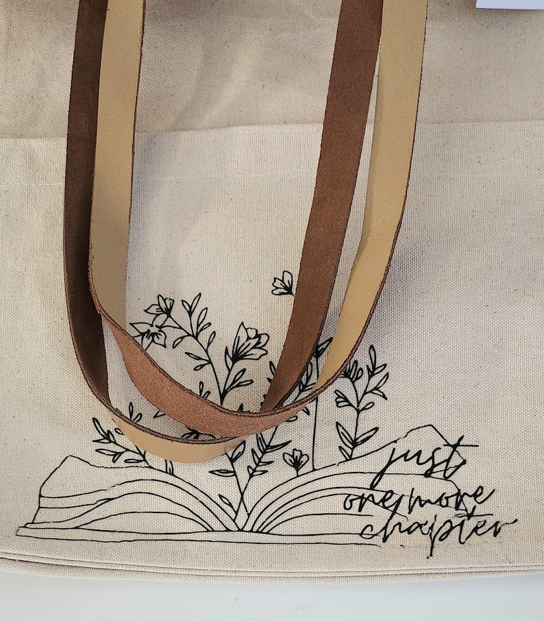 May include: Cream tote bag with brown and tan leather straps. The bag has a black line drawing of an open book with floral accents and the text "just one more chapter."