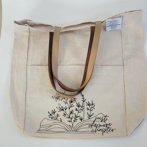 May include: A natural canvas tote bag with brown leather handles. The bag features a black line drawing of an open book with floral accents and the text "just one more chapter."