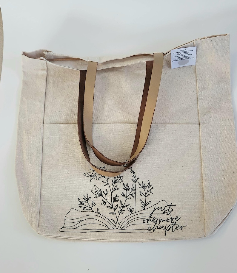 May include: A natural canvas tote bag with brown leather handles. The bag features a black line drawing of an open book with floral accents and the text "just one more chapter."