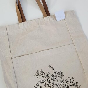 May include: Cream canvas tote bag with brown leather handles. The bag has a front pocket and a black floral and book design with the text "just one more chapter."