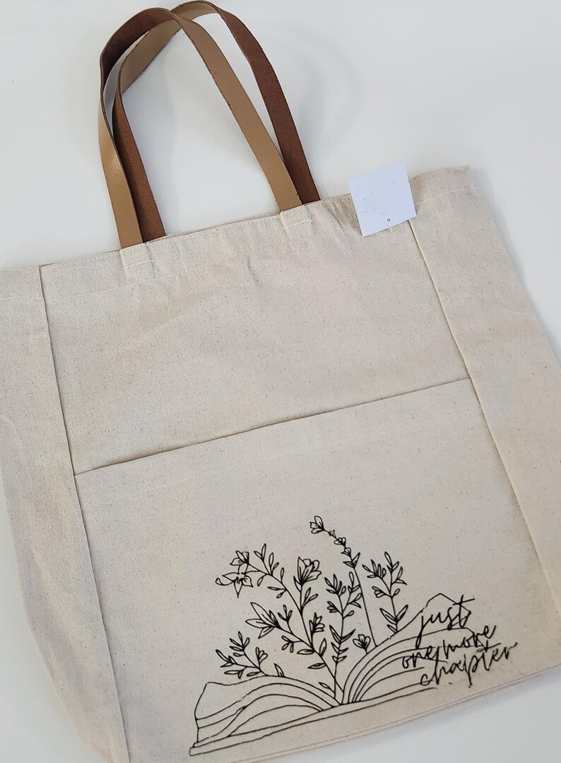 May include: Cream canvas tote bag with brown leather handles. The bag has a front pocket and a black floral and book design with the text "just one more chapter."
