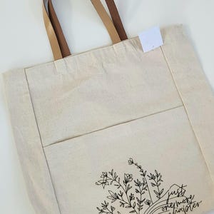 May include: Cream canvas tote bag with brown leather handles. The bag has a front pocket and a black floral design with the text "just one more chapter". A stylish bag for carrying books and other items. The bag is made of canvas.