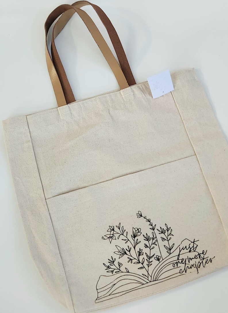 May include: Cream canvas tote bag with brown leather handles. The bag has a front pocket and a black floral design with the text "just one more chapter". A stylish bag for carrying books and other items. The bag is made of canvas.