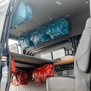 May include: The interior of a van with a built-in counter and several blue and red duffel bags hanging from the ceiling and side walls.