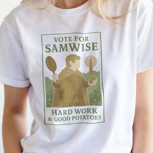 Op de afbeelding: Wit T-shirt met een afbeelding met de tekst "VOTE FOR SAMWISE" boven een illustratie van een persoon die een koekenpan en een lantaarn vasthoudt. De tekst "HARD WORK & GOOD POTATOES" staat eronder. Het shirt heeft een ronde hals.