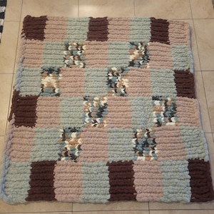May include: A handmade, chunky knit blanket in a patchwork design. The blanket features alternating squares of brown, light blue, and pink. Some squares have a speckled pattern with hints of blue, brown, and cream. The blanket is made of a soft, plush material.