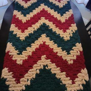 May include: A rectangular hand-knitted blanket featuring a repeating chevron pattern in deep forest green, dark red, and cream yarn. The chunky knit texture is consistent throughout the piece, showcasing a thick, soft, and looped yarn construction.