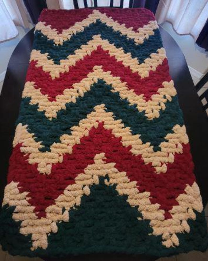 May include: A rectangular hand-knitted blanket featuring a repeating chevron pattern in deep forest green, dark red, and cream yarn. The chunky knit texture is consistent throughout the piece, showcasing a thick, soft, and looped yarn construction.