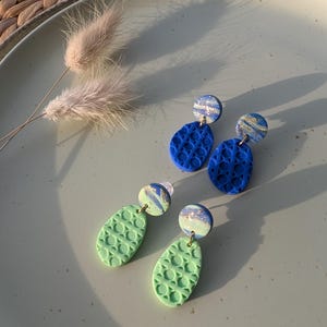 Könnte beinhalten: Zwei Paar handgefertigte Polymer-Ton-Ohrringe. Ein Paar ist leuchtend blau mit einem strukturierten Muster, das andere hellgrün mit einem ähnlichen Design. Jeder Ohrring hat ein kleines, marmoriertes Akzentstück mit Goldflocken.