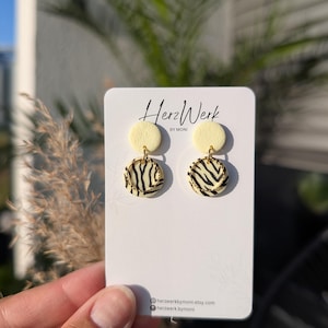 Könnte beinhalten: Handgefertigte Ohrringe mit einem cremefarbenen oberen Kreis und einem unteren Kreis mit Zebra-Print. Die Ohrringe werden auf einer weißen Karte mit dem Text "HerzWerk by MONI" und Social-Media-Details präsentiert. Gehalten von einer Hand.