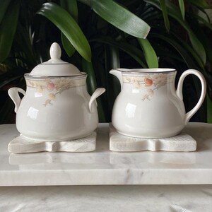 May include: A white sugar bowl and creamer set with a floral design. The set is on a white marble surface.