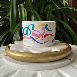 May include: White ceramic planter with a colourful abstract design featuring swirls and lines in pink, blue, green, yellow, and red. The planter is sitting on a white marble and gold tray.