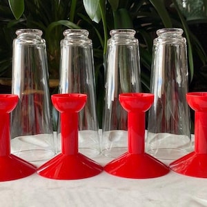 May include: Six clear glass cups with red plastic bases. The cups are tall and cylindrical, and the bases are shaped like a wide, flat disc with a raised center.