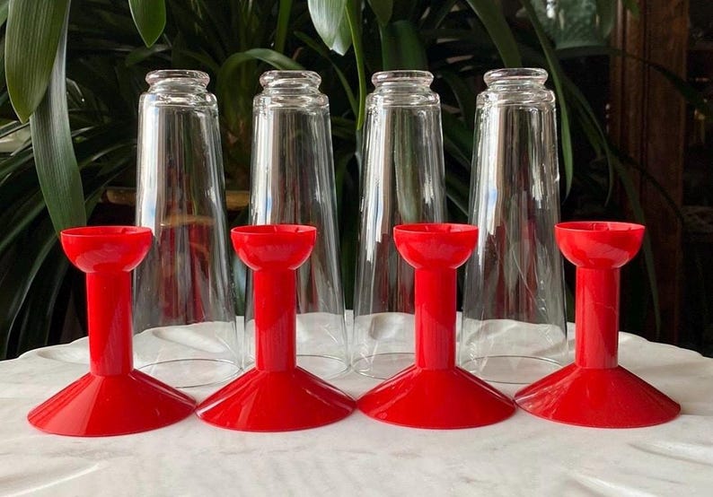 May include: Six clear glass cups with red plastic bases. The cups are tall and cylindrical, and the bases are shaped like a wide, flat disc with a raised center.