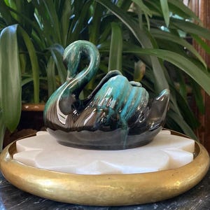 Puede incluir: Una figurita de cisne de cerámica con un esmalte negro y verde. El cisne está sentado sobre una base de mármol blanco con un borde dorado.