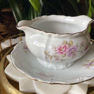 May include: White porcelain gravy boat and saucer set. The gravy boat has a scalloped rim with gold trim and is decorated with pink and purple floral patterns. The saucer also has gold trim and matching floral designs.