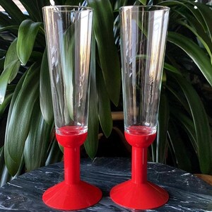 May include: Two clear glass champagne flutes with red plastic bases. The flutes have a delicate etched design.