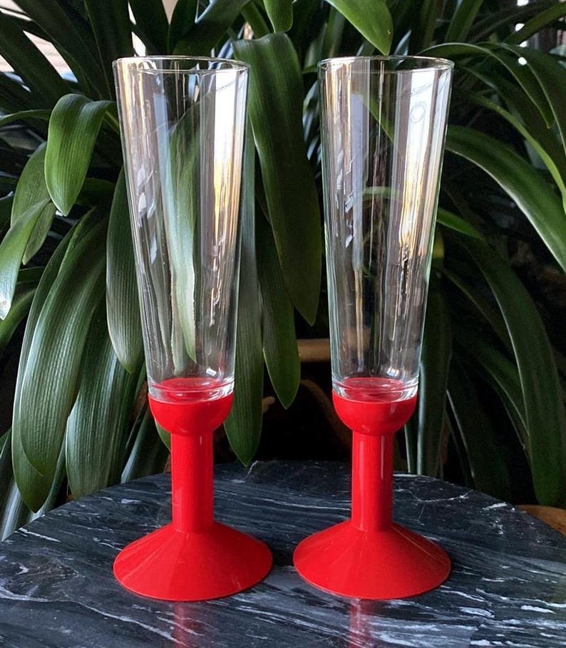 May include: Two clear glass champagne flutes with red plastic bases. The flutes have a delicate etched design.