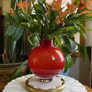 Peut inclure: Un vase rond en verre rouge vif avec une large ouverture contient un bouquet de fleurs orange et jaunes. Le vase est posé sur une surface en marbre blanc avec une base en métal doré.