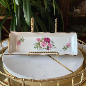 May include: A white rectangular ceramic dish with a gold trim and a floral design featuring pink and red roses. The dish is on a gold stand.
