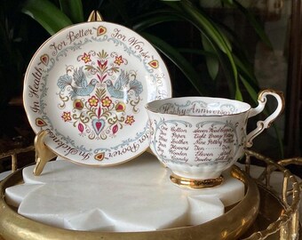 May include: A vintage teacup and saucer set. The white porcelain saucer features two blue birds, floral designs, and the text "For Better, For Worse, In Sickness, In Health, For Poorer". The teacup has gold trim and the text "Happy Anniversary".