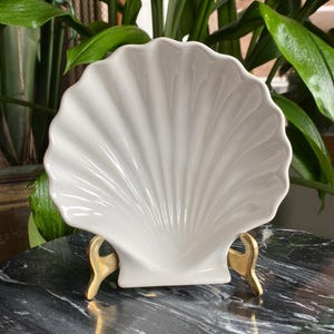 May include: A white ceramic seashell dish with a gold metal stand. The dish is shaped like a seashell and has a smooth, glossy finish.