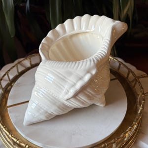 May include: A cream-coloured ceramic seashell-shaped planter. The planter has a textured surface with ribbed detailing and a ruffled edge. It sits on a white marble-like surface within a gold-toned tray, creating a coastal or nautical theme.