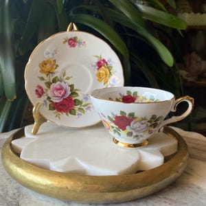 May include: A white teacup and saucer with a floral pattern featuring red, pink, yellow, and white roses. The cup and saucer have a gold trim.