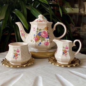 Puede incluir: Un juego de té floral vintage con tetera, jarra de crema y azucarero. El juego es blanco con un patrón floral en tonos de rosa, azul y amarillo. El juego está en una bandeja de metal dorado.