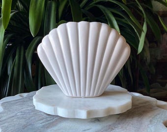 May include: A white, seashell-shaped decorative object with vertical ridges, resting on a white, scalloped-edge marble base. The background features lush green foliage, creating a natural, coastal aesthetic. The object is likely ceramic or porcelain.
