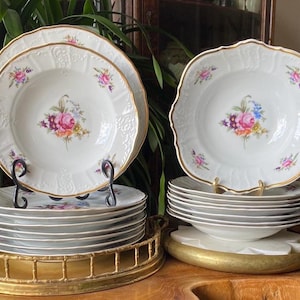 May include: A set of white porcelain plates with gold rims and floral center designs. The plates are stacked on a gold tray and plate stands. The floral design features pink, purple, and yellow flowers.