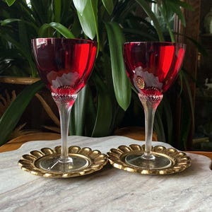May include: Two red wine glasses with gold accents on the base, sitting on gold metal coasters.