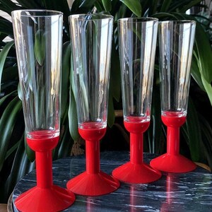 May include: Set of four clear glass champagne flutes with red plastic bases. The flutes are tall and slender with a wide bowl. The bases are a bright red color and have a flared shape.