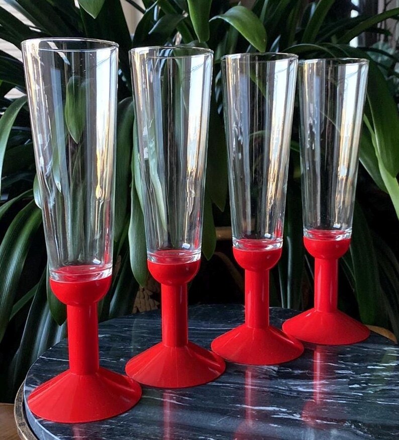 May include: Set of four clear glass champagne flutes with red plastic bases. The flutes are tall and slender with a wide bowl. The bases are a bright red color and have a flared shape.