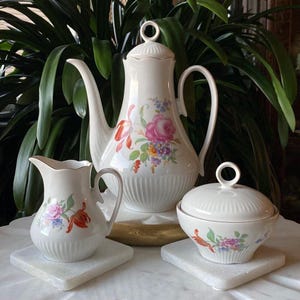 Puede incluir: Un juego de té de porcelana blanca con diseños florales. El juego incluye una tetera, una jarra de crema y un azucarero con tapa. Las flores son rosas, rojas y amarillas.