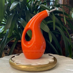 Może przedstawiać: Pomarańczowa ceramiczna butelka na olej i ocet z wylewką ze stali nierdzewnej. Butelka ma unikalny, nowoczesny design z otworem po boku, ułatwiającym nalewanie.
