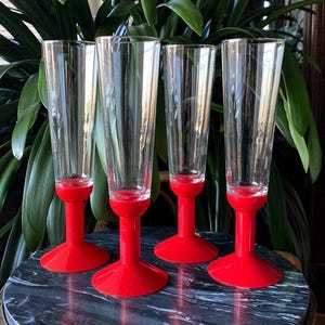 May include: Four clear glass champagne flutes with red plastic stems and bases. The flutes are arranged in a row on a black surface.