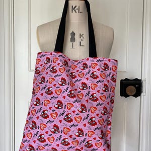 May include: Pink tote bag with black handles, featuring a repeating pattern of red hearts, bats, and the word "LOVE" in a stylized font. The bag is displayed on a mannequin.
