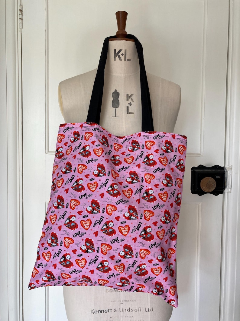 May include: Pink tote bag with black handles, featuring a repeating pattern of red hearts, bats, and the word "LOVE" in a stylized font. The bag is displayed on a mannequin.