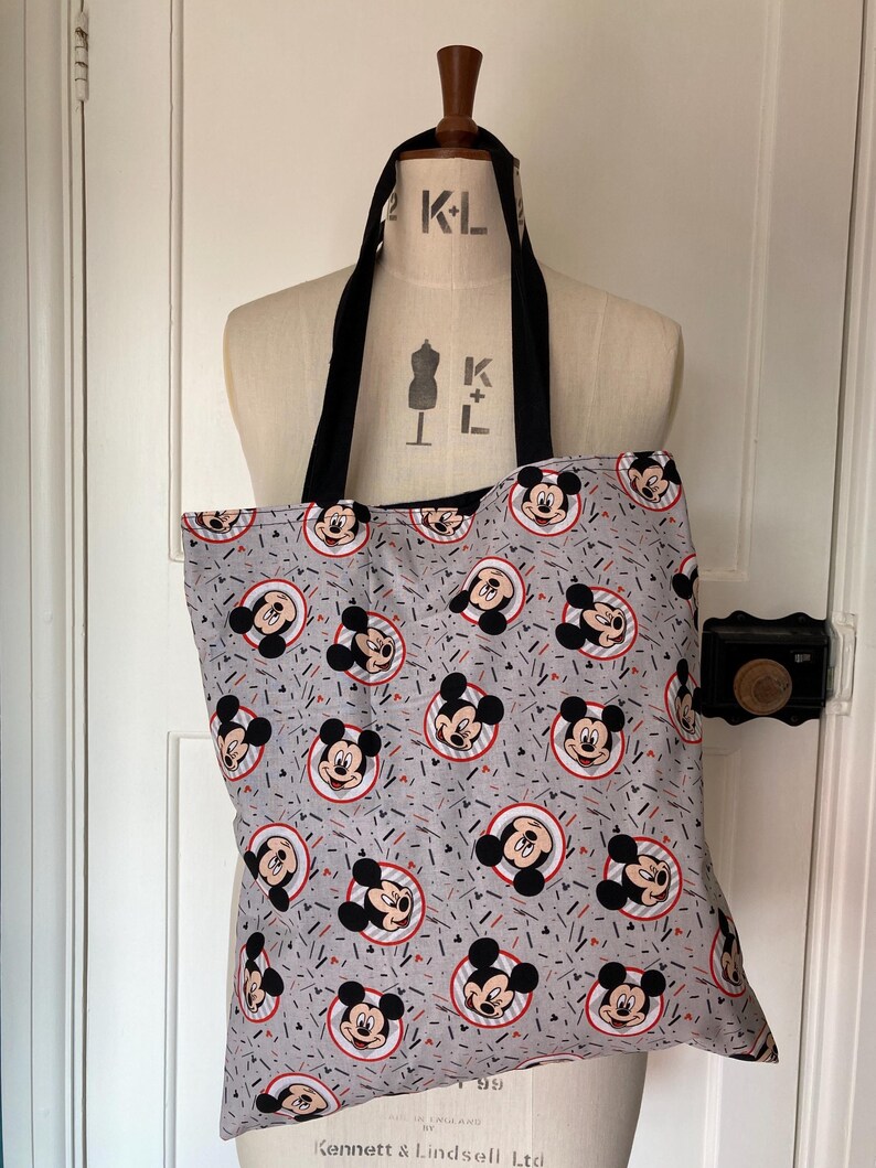 May include: A gray tote bag with a black handle and a repeating pattern of Mickey Mouse heads in red, black, and white on a gray background with red and white confetti.