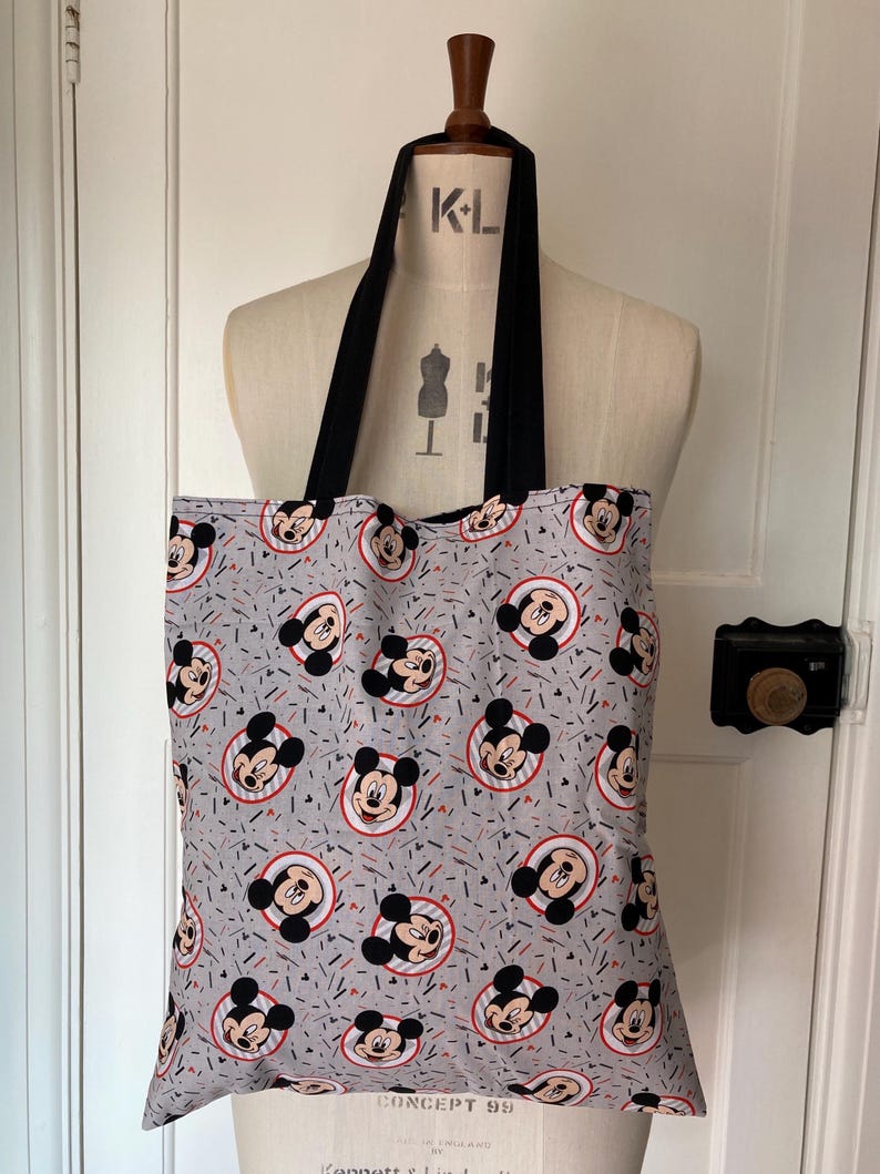 May include: A gray tote bag with black straps featuring a repeating pattern of Mickey Mouse heads in red, black, and white on a gray background with red and blue confetti.