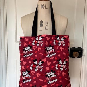 May include: Red tote bag with a black handle featuring a repeating pattern of Mickey Mouse and Minnie Mouse holding hands with hearts and flowers.