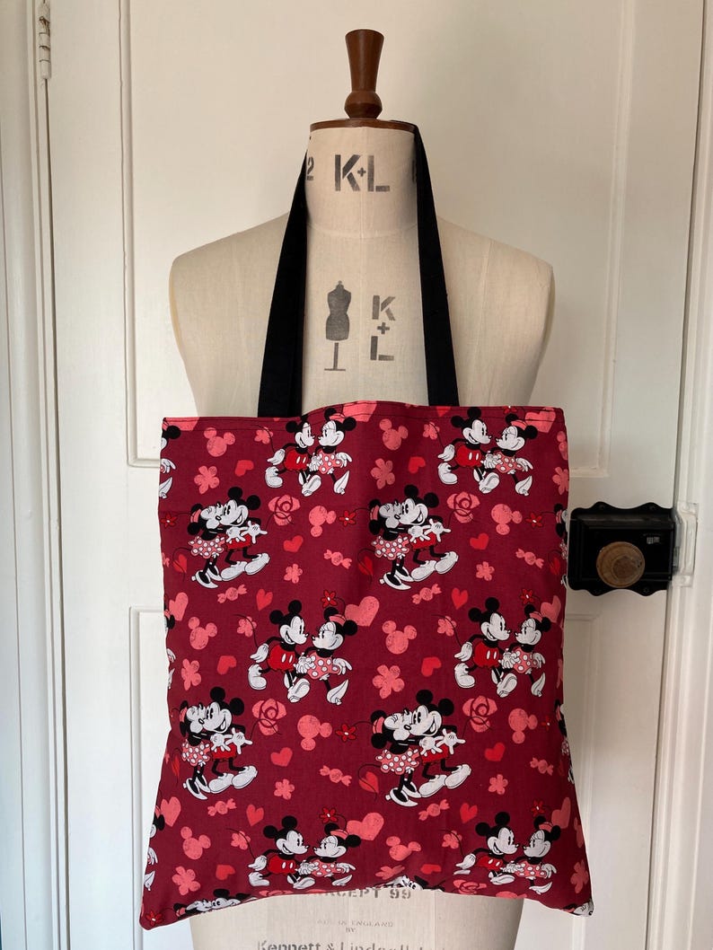 May include: Red tote bag with a black handle featuring a repeating pattern of Mickey Mouse and Minnie Mouse holding hands with hearts and flowers.