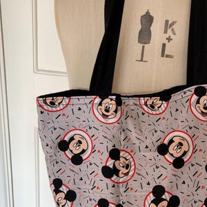 May include: A gray tote bag with black straps and a repeating pattern of Mickey Mouse heads in red circles on a gray background. The background also has red and black lines.