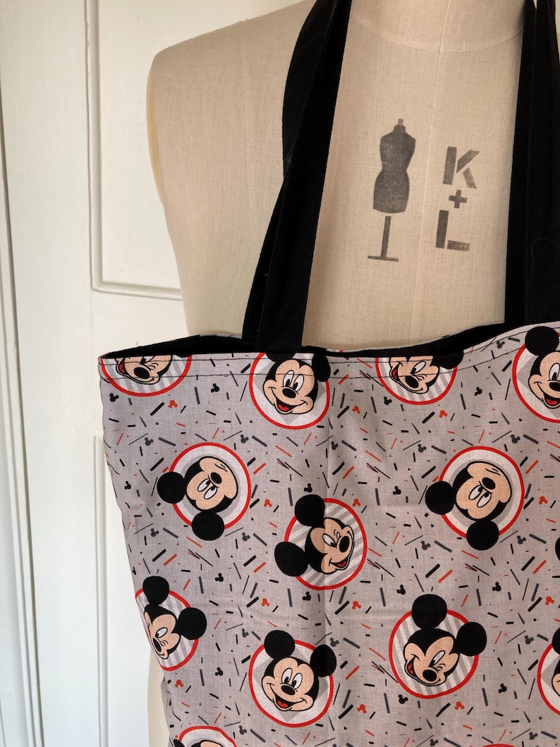 May include: A gray tote bag with black straps and a repeating pattern of Mickey Mouse heads in red circles on a gray background. The background also has red and black lines.