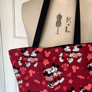 May include: A red tote bag with a black handle. The bag is decorated with a repeating pattern of Mickey Mouse and Minnie Mouse in a romantic pose. The pattern also includes hearts and roses.